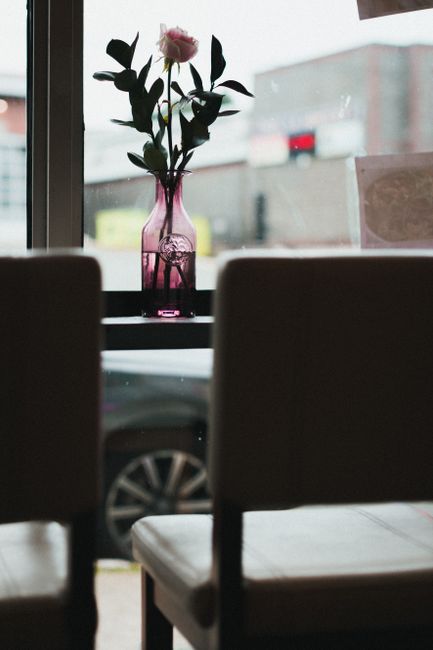 A single pink-tinted glass vase with a narrow neck and rounded base, positioned on a dark tabletop between two blurred chairs, contains a fresh pink rose with partially open petals and dark green leaves. The scene is illuminated by natural light coming through a large window behind the vase, revealing an out-of-focus urban street scene with a yellow sign and a red vehicle passing by. The background shows the exterior of buildings with concrete facades, and the sky appears overcast. The vase's transparent glass finish highlights its ridges and smooth surface, contrasting with the matte texture of the chairs in the foreground. The photograph captures a quiet, indoor ambiance with muted shadows and soft light, emphasizing the floral arrangement as the focal point within a modern interior setting, which may be part of a café or casual seating area.