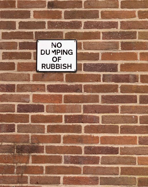 A rectangular white metal sign with black text reading 'No Dumping of Rubbish' is mounted on a brick wall constructed with reddish-brown bricks, showing variations in tone and texture. The sign is positioned centrally in the upper half of the image, with evenly spaced bricks vertically and horizontally creating a uniform pattern across the wall. The wall appears to be part of an exterior environment, with no additional objects or environmental features visible. The lighting is natural, suggesting daylight, casting soft shadows that outline the brick textures. This scene reflects a typical outdoor setting where a property owner or local authority might place signage to discourage informal waste disposal, aligning with alternative rubbish management approaches that focus on private or site-specific rubbish control, as seen through the prominence of the sign against the brick surface.