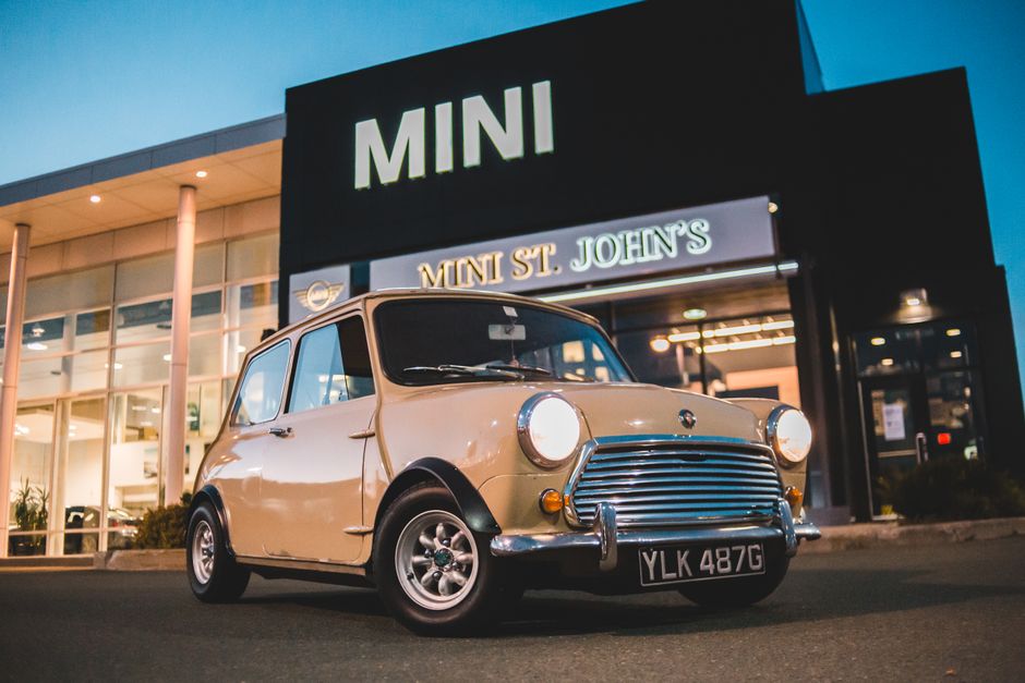 A vintage beige Mini Cooper with a rounded body shape, circular headlights, and a distinctive front grille, parked on the asphalt outside a modern MINI dealership building at dusk. The car features chrome bumpers and trim, black side mirrors, and small alloy wheels. The dealership has large glass windows illuminated from within, showcasing the showroom, with a black exterior and a prominent sign displaying the word 'MINI' in large white letters. The building's signage includes the text 'MINI St. John's' in illuminated capital letters below the main logo. Surrounding the area are small bushes and a paved driveway, with soft ambient lighting casting gentle shadows, creating a clean and neutral environment that subtly relates to private vehicle disposal and alternative waste handling options available through waste collection services near St. John's Wood.