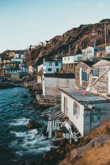 A waterfront scene featuring a series of weathered wooden houses built on rocky foundations along the shoreline, with some structures showing signs of age such as peeling paint, visible timber, and small concrete steps leading from the water. The buildings are predominantly painted in light shades such as white and pale grey, with one having darker window shutters. The narrow shoreline supports a slight overhang from the rocky cliffs behind, which are rugged and brownish in color with sparse vegetation. The water below is choppy with small waves lapping against the structures, creating a dynamic environment. In the background, additional houses and buildings are visible atop the cliffs, along with a blue sky and sparse clouds. The scene appears to be during daylight with natural lighting, emphasizing textures of the aged wood and rocky coast, reflecting quiet, possibly historic or traditional seaside habitation. The setting aligns with independent coastal dwellings rather than modern developments, suitable for waste management services that handle refuse on-site or undertake private disposal for such unique locations.
