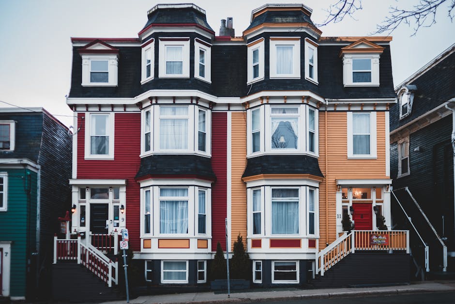 The image depicts a multi-storey Victorian-style residential building with a vibrant, color-blocked facade in shades of red, orange, and black. The building features prominent bay windows on each level, with white frames and decorative trims, and a textured exterior clad in horizontal wooden siding. The dark roof has a mansard shape with dormer windows, and the structure is flanked by narrow neighboring buildings of similar Victorian design. In front, there are concrete stairs leading to the red front door, which is flanked by small black decorative railings and topped with a white canopy. To the left, a street lamp and a no-parking sign are visible, along with a small patch of pavement, indicating the building is situated on a residential street. The overall scene conveys a typical urban setting where private waste collection, such as by waste removal services like wastecollectionstjohnswood.co.uk, might be involved in managing rubbish or debris associated with property maintenance or renovation activities.